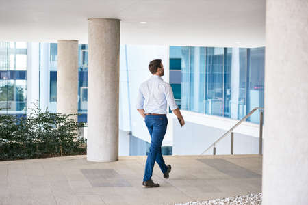 Rear view young businessman walking with a digital tablet along a footpath outside of a modern...