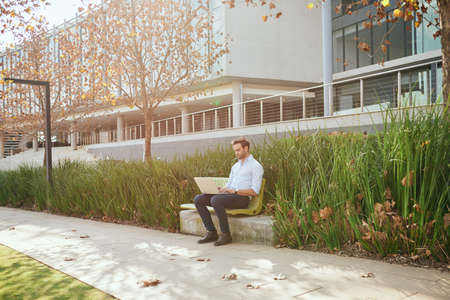 Young businessman working on a laptop while sitting on a bench outside of a modern office complexの写真素材