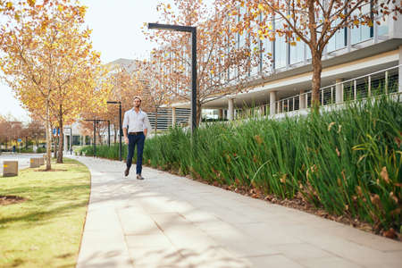 Casually dressed young businessman walking outside on a footpath by an office complex in autumn