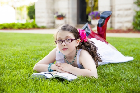 Beautiful young hispanic girl in glasses, white dress, and red boots, reads her book outside on the grassの写真素材