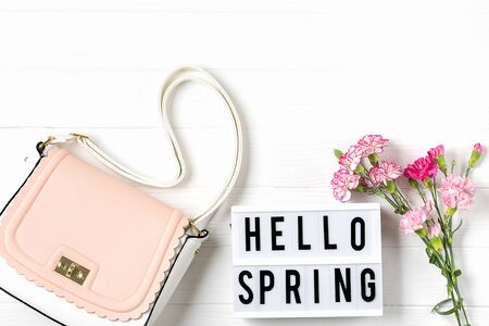 Bouquet of pink carnations flowers, lightbox with text Hello Spring, womans handbag on white wooden...