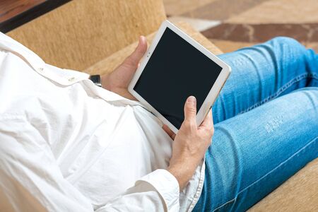 young man in white shirt and blue jeans working and looking on tablet with black screen in home office on cozy beige sofa Stay at home, online work, distance learning concept Top view Mock up.の写真素材