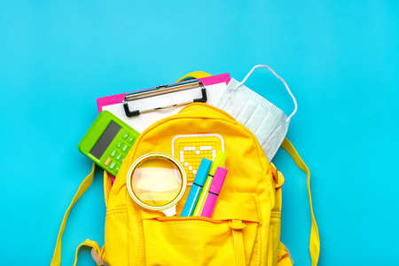 Back to school, education concept Yellow backpack with school supplies, protective medical mask, calculator, scissors isolated on blue background. Top view Copy space Flat lay.の写真素材