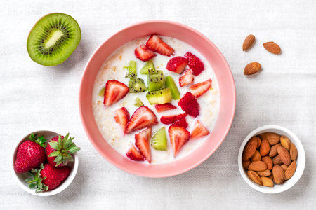 oatmeal porridge with slices of kiwi, strawberries, almonds in pink bowl, spoon on white tablecloth Top view Flat lay Vegan food concept Healthy breakfastの写真素材