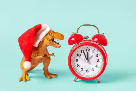 Dinosaur Rex in red Santa Claus hat and alarm clock on blue background New Years Eve or Christmas...