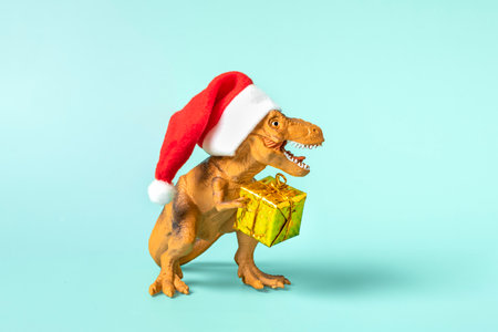 Dinosaur Rex in red Santa Claus hat holds golden gift box in its paws on blue background New Years Eve or Christmas Eve Art holiday card Creative idea for Merry xmas concept.の写真素材
