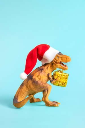 Dinosaur Rex in red Santa Claus hat holds golden gift box in its paws on blue background New Years Eve or Christmas Eve Art holiday card Creative idea for Merry xmas concept.の写真素材