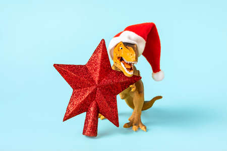 Dinosaur Rex in red Santa Claus hat holds star in its paws on blue background New Years Eve or...