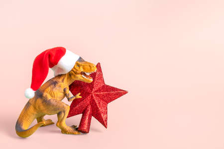 Dinosaur Rex in red Santa Claus hat holds star in its paws on pink background New Years Eve or Christmas Eve Art holiday card Creative idea for Merry xmas concept.の写真素材