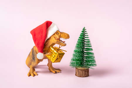 dinosaur Rex in red Santa Claus hat holds golden gift box in its paws and tree on pink background New Years Eve or Christmas Eve Art holiday card Creative idea for Merry xmas concept.の写真素材