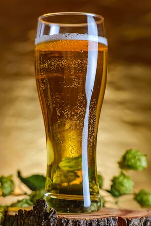 Amber beer with a sprig of hops on a wooden Board. Foam hop drink. Foamy beer in a glass on a wooden stand, gold blurred backgroundの写真素材