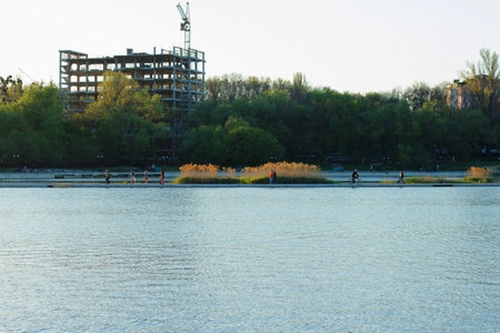 Construction of a multi-storey building in park area, where people walk, there is a lake and green trees. Landscapeの写真素材