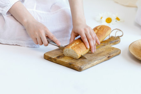 Girl cuts a loaf of bread with a knife on a wooden baking sheet on the floor. Part of the body. Focus on handsの写真素材