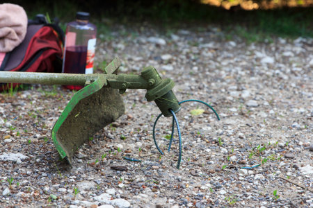 Green grass trimmer tool lie on gravel ground with blurred background of red bag and bottle, outdoor...