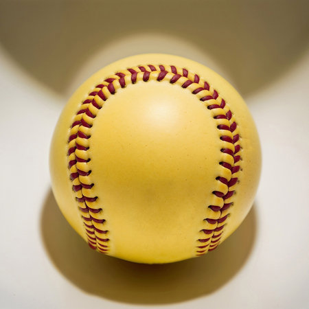 A close-up of a yellow softball with red stitching, placed on a light background. Ball is round and smooth, typical for the sport of softballの素材