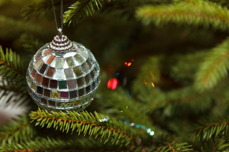 A shiny disco ball ornament hangs from a Christmas tree branch. Ornament reflects light, creating a festive atmosphere with colorful bokeh in backgroundの写真素材