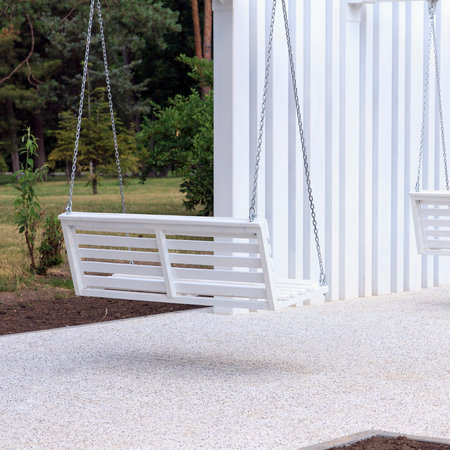 Modern outdoor space featuring white swings under a minimalist structure. The area is surrounded by greenery and flower beds, creating a serene environmentの写真素材