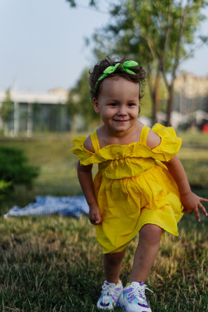 A curly-haired toddler is seen smiling brightly in a sunny outdoor setting. She is dressed in a yellow dress and sneakers, with a green bow in her hair. The background shows a grassy area with trees and a blurred view of a building.の写真素材