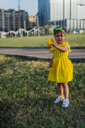 A young girl in a park, holding a yellow ball, and wearing a yellow dress and green headband.の写真素材
