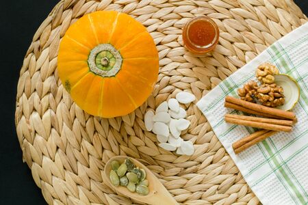 Small pumpkin with seeds, peeled seeds in wooden spoon,  little glass can of honey, walnuts and cinnamon sticks on a circle mat/napkin made of water hyacinth on a black background and green-white checkered waffle towelの写真素材