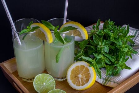 Homemade citrus lemonade in glasses with straws with mint leaves and a bunch of mint, lemons, limes on a bamboo tray and whte towel on a black background. Close upの写真素材