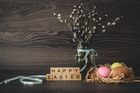 The inscription  happy Easter, made of light wooden cubes, painted eggs in the nest, willow twigs with seals on a dark wooden backgroundの写真素材