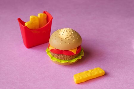 Plastic burger, salad, tomato, frying potatoes on a pink background. Horizontal orientation....