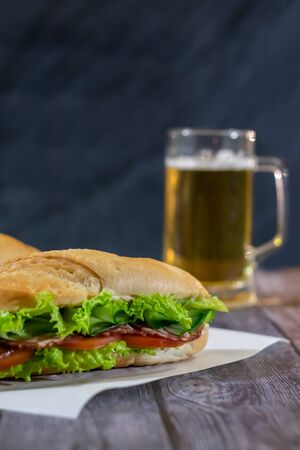 Two fresh submarine sandwiches with sausage, cheese, bacon, tomatoes, lettuce, cucumbers and onions on a dark wooden background. Mug of beerの写真素材