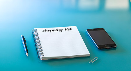 Blank notebook with a shopping list, pen, and smartphone on a blue table, providing an organized workspace for planning purchases and daily tasksの素材