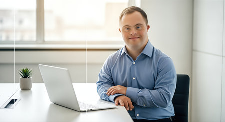 Young man with Down syndrome is seated at a modern desk in a well-lit office, engaging with a laptop, reflecting positivity and social interactionの素材
