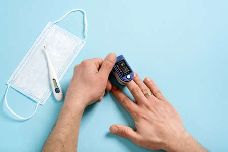 Human male hands pulse oximeter used to measure pulse rate and oxygen levels with medical blue background with copy space. Top view. Medical concept protective medical mask, electronic thermometerの素材 [FY310172520779]