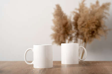 Mockup two white mugs on a wooden table top with pampas decor in a Scandinavian interior, boho style. Template, layout for your design, advertising, logo with copy space. Cups light beige backgroundの写真素材
