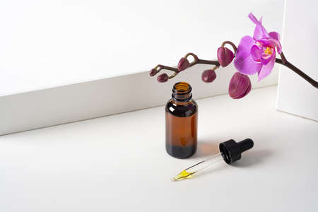 Mockup cosmetic face serum, oil brown glass bottle with a pipette on a stylish minimalist white background with geometric decor and orchid flowers. Female beauty concept, empty template. Copy spaceの写真素材