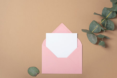 Real photo. Pink envelope square invitation card mockup with a eucalyptus branch. Top view with copy space, pastel beige background. Template for branding and advertisingの写真素材
