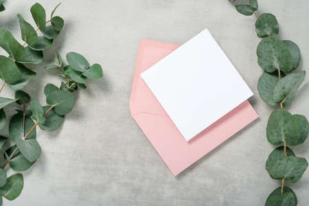 Real photo. Pink envelope square invitation card mockup with a eucalyptus branch. Top view with copy space, light gray concrete background. Template for branding and advertisingの写真素材