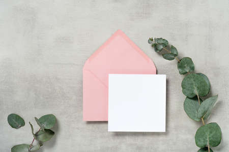 Real photo. Pink envelope square invitation card mockup with a eucalyptus branch. Top view with copy space, light gray concrete background. Template for branding and advertisingの写真素材