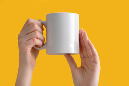 mockup ceramic white coffe cup or mug in female hands on yellow background with copy space. Blank template for your design, branding, business. Woman in shirt casual style. Real photo.の写真素材
