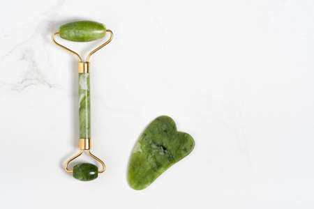 Jade roller for face massage. Green gua sha facial massager tools on the marble white background with copy space flat lay. Anti age, lifting and toning treatment, acupressure.の写真素材