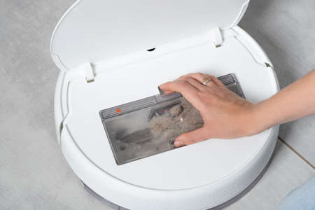 A woman opens the lid of a white robotic vacuum cleaner to reveal a transparent plastic container with small debris and cat pet hair on a concrete tile floor of a house close-up.の写真素材