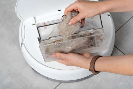 A woman opens the lid of a white robotic vacuum cleaner to reveal a transparent plastic container with small debris and cat pet hair on a concrete tile floor of a house close-up.の写真素材