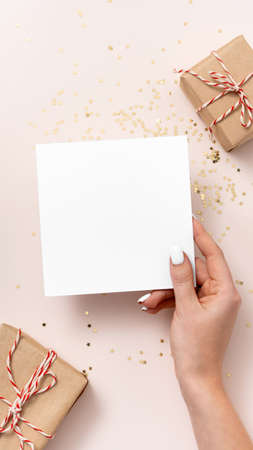 Female hand holds blank square paper mockup, golden stars confetti, gift boxes on beige background. Flat lay, top view, copy space, minimalist. Christmas and New Year conceptの写真素材