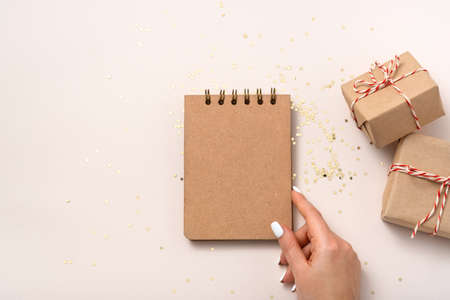Female hand hold notebook blank paper mockup, golden stars confetti, gift boxes on beige background. Flat lay, top view, copy space, minimalist. Christmas New year composition.の写真素材