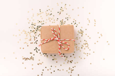 Gift box in craft paper with white-red New Year's ribbon and gold confettistars on a beige...