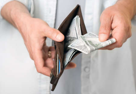 A man in casual clothes holds a male brown wallet with money and hundred dollar bills in his hands....