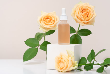 Mockup cosmetics serum with a pipette in a transparent frosted glass and orange liquid. Facial peeling with lactic Aha acid on a white cubic pedestal, beige background with rosesの写真素材