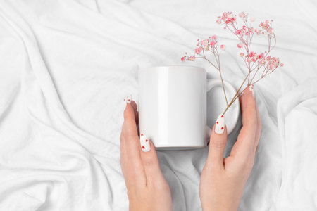 Women's hands hold a mockup of a white empty mug on a textile background, a cup for your design and  close-up. White manicure nails with red hearts for Valentine's Day.の写真素材