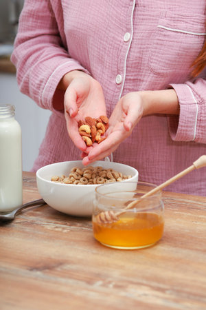 Woman female hands prepare a quick breakfast of dry round corn flakes balls, honey, milk on a kitchen. Nuts, almonds and cashewsの写真素材