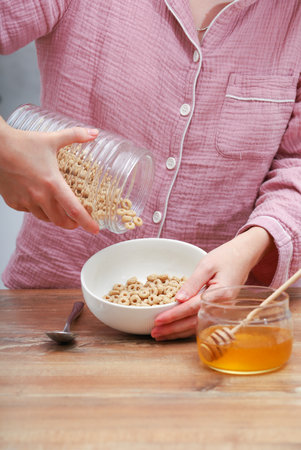 Woman female hands prepare a quick breakfast of dry round corn flakes balls, honey, milk on a kitchen.の写真素材