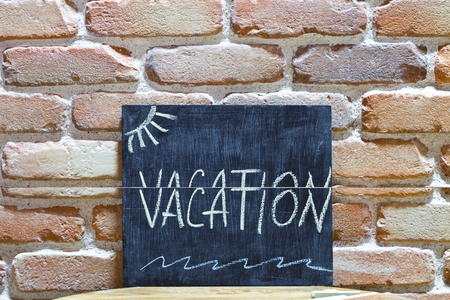 Chalk board with the word VACATION drown by hand and chalks on wooden table on brick wall background. Symbol for presentation.の写真素材
