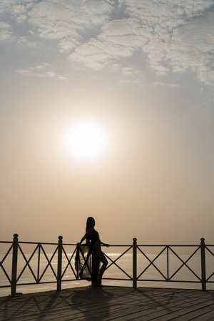 A girl in a swimsuit and a cloak is standing on the pier and meets the dawn.の写真素材